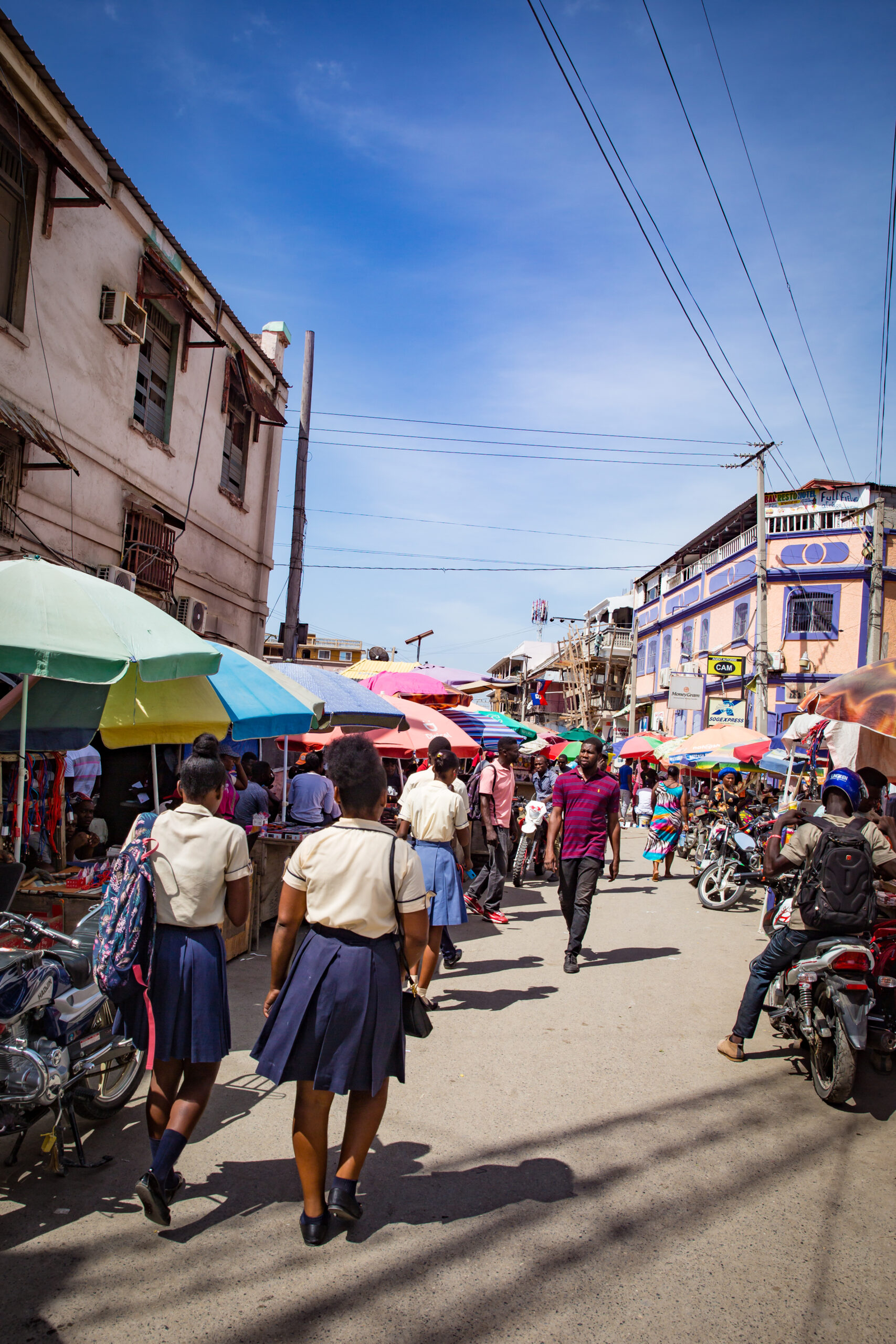 A crowded street market with colorful umbrellas, people walking, and various shops and buildings visible.