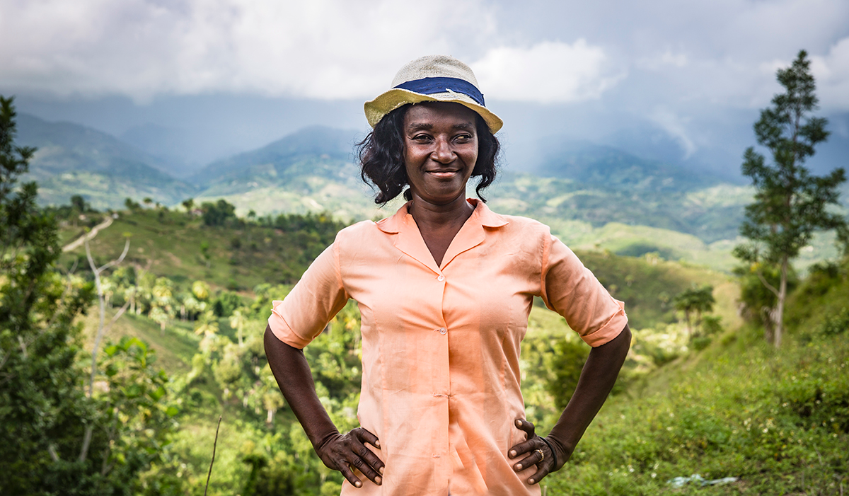 A smiling woman wearing a peach-colored shirt and hat stands amidst lush, mountainous greenery.