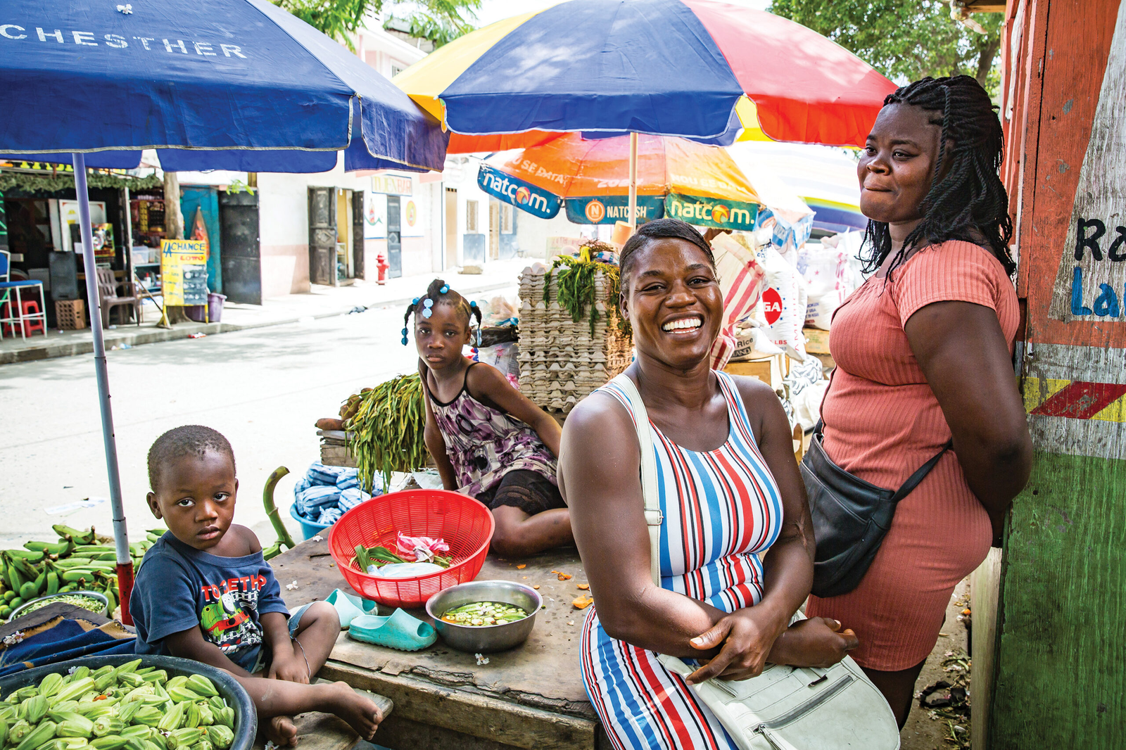 A group of people, including a smiling woman and children, selling fresh produce at a market stall.
