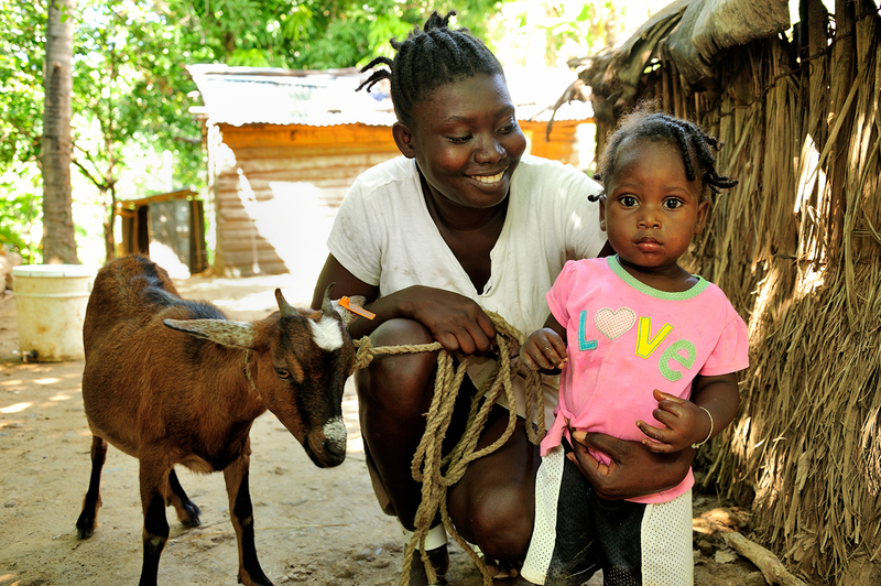A smiling woman holds a young child, who wears a pink shirt with the word "Love", as they stand next to a small brown goat.
